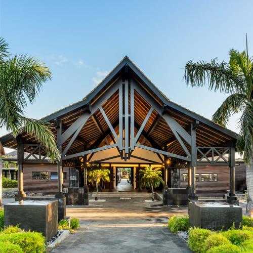 Moderner tropischer Hoteleingang mit großem, hölzernem A-förmigem Dach, umgeben von Palmen und üppigem Grün bei klarem blauem Himmel.