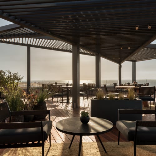 Außenbereich auf dem Dach mit modernen Stühlen und Tischen unter einem lamellierten Pergola, mit Blick auf ein Gewässer bei Sonnenuntergang und sanftem Naturlicht.