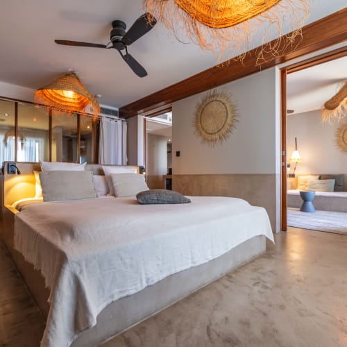 Modernes Schlafzimmer mit einem großen Bett, das mit weißen Bettwäsche bedeckt ist, Deckenventilatoren und gewebten Lampenschirmen, durch eine große offene Tür ist ein angrenzender Raum sichtbar.