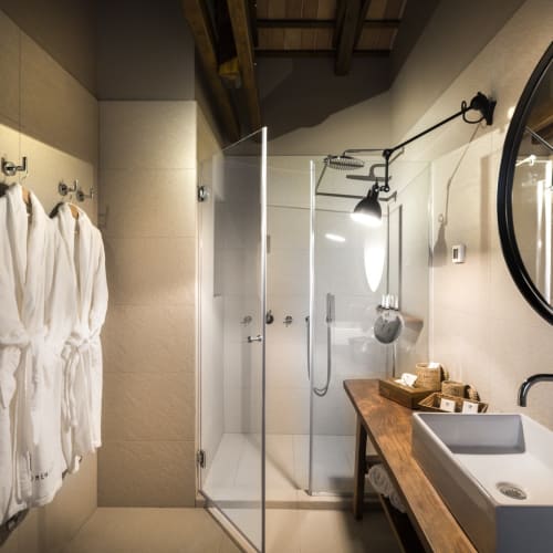 A modern bathroom with a glass shower partition, white bathrobes hanging on hooks, a wooden sink counter, and a round mirror on the wall.