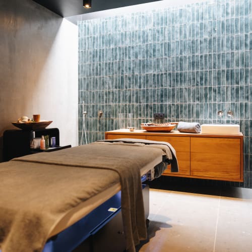 Ein moderner Massageraum mit einer mit einem beigen Handtuch bedeckten Massageliege, einem Holzschrank mit gerolltem Handtuch und Dekorationsgegenständen sowie einer Wand mit vertikal angeordneten grünen Fliesen.