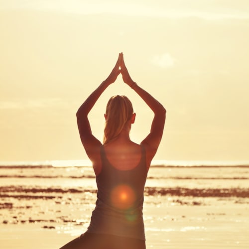Eine Frau macht die Baumstellung im Yoga am Meer während eines goldenen Sonnenuntergangs, als Silhouette vor dem hellen Himmel und Wasser.