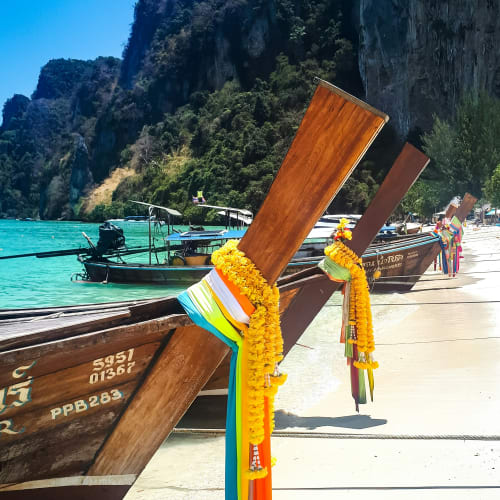 Traditionelle thailändische Langboot-Boote mit farbigen Blumengirlanden am Bug, am sandigen Strand vor türkisfarbenem Wasser und bewachsenen Felsen im Hintergrund bei klarem Himmel.