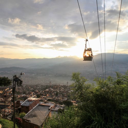 Eine Seilbahn, die über eine Stadtlandschaft mit grüner Vegetation und Bergen im Hintergrund bei Sonnenuntergang fährt.