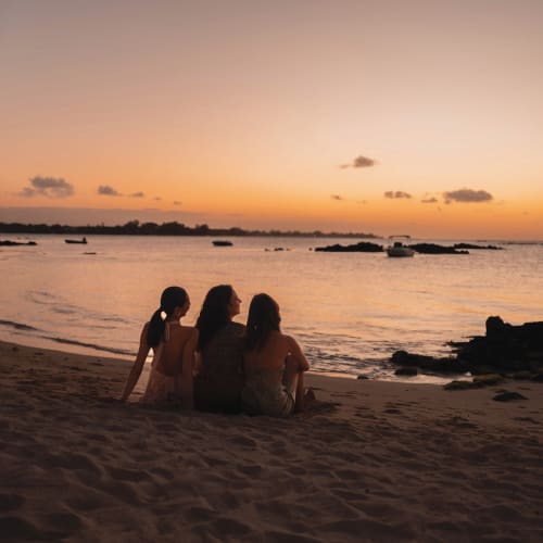 Drei Frauen sitzen am Strand während eines Sonnenuntergangs und blicken auf das Wasser und Boote auf dem Meer.