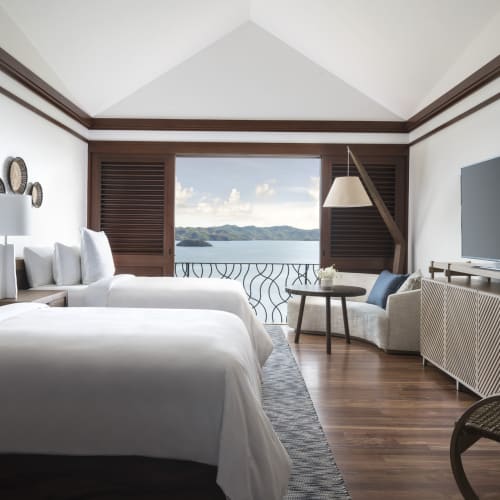 Luxurious hotel room with two twin beds, a sitting area, and a large window overlooking the ocean and islands.