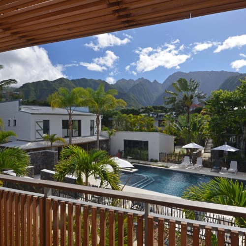Blick von einem hölzernen Balkon auf einen ruhigen Swimmingpool, umgeben von tropischen Palmen und modernen weißen Gebäuden, mit grünen Bergen unter einem teils bewölkten blauen Himmel im Hintergrund.