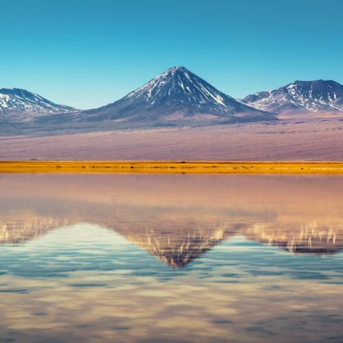A panoramic view of the Atacama Desert with snow-capped mountains reflected in a lake.