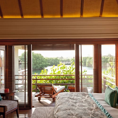 Luxuriöses Schlafzimmer mit Balkon, der auf üppiges Grün blickt, mit großem Bett, Holzmöbeln und warmem Licht.