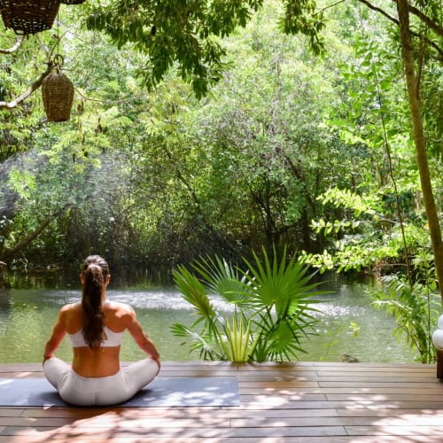 A woman practicing yoga on a mat near a tranquil water body surrounded by lush green trees in a tropical setting.