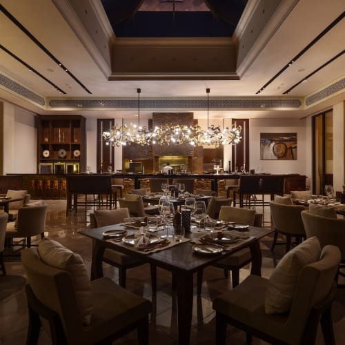 Schummrig beleuchtetes Interieur eines modernen Restaurants mit eleganten Holztischen, die zum Essen gedeckt sind, umgeben von gepolsterten Stühlen und beleuchtet von einem zeitgenössischen Kronleuchter über dem Barbereich.