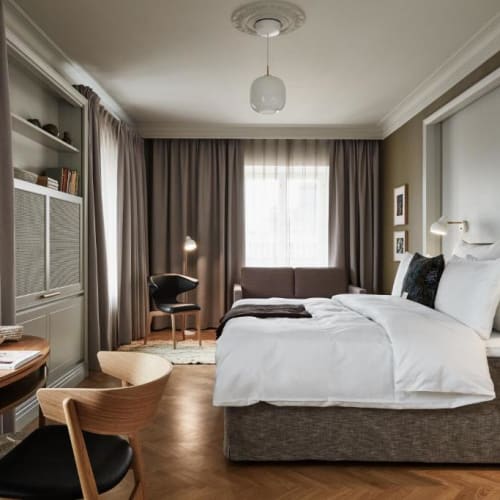 Modernes und gemütliches Hotelzimmer mit großem Bett, Holzboden, Schreibtisch mit Stuhl, Regalen und Fenster mit Vorhängen, das natürliches Licht hereinlässt.