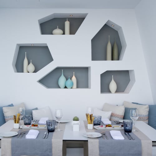 Ein modernes Esszimmer mit einem weißen Tisch, der für vier Personen gedeckt ist, mit Glaswaren, Tellern und Geschirr, vor einer blauen gepolsterten Bank und geometrischen Wandregalen mit Vasen.