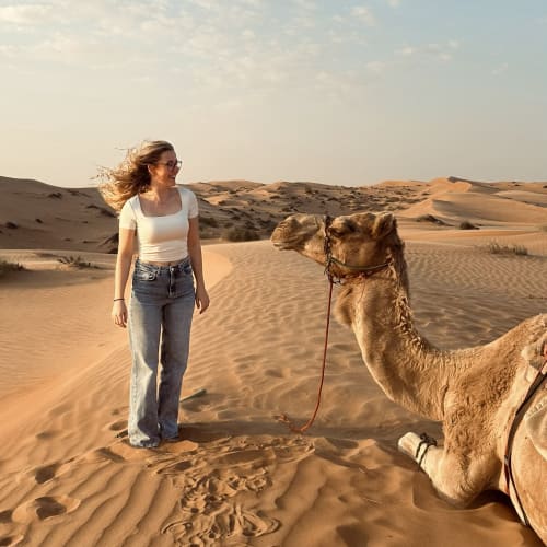 Eine Frau mit Brille, weißem Oberteil und Jeans steht auf Sanddünen und lächelt ein auf dem Wüstensand sitzendes Kamel mit Zaumzeug und Zügeln an.