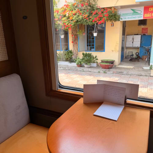 A cozy train compartment with a wooden table and cushioned seat next to a window showing a street view with a yellow building, potted plants, and a flowering tree outside.