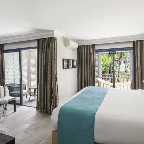 Helles Junior-Suitenhotelzimmer mit großem Bett mit weißer Bettwäsche und türkisfarbenem Läufer, gemütlicher Sitzecke mit Sofa und Tisch sowie zwei Schiebetüren zu Balkonen mit Außenblick und Hängematte.