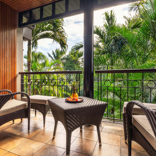 A balcony with wicker chairs and a small table with a pineapple centerpiece, surrounded by lush tropical palm trees under a partly cloudy sky.