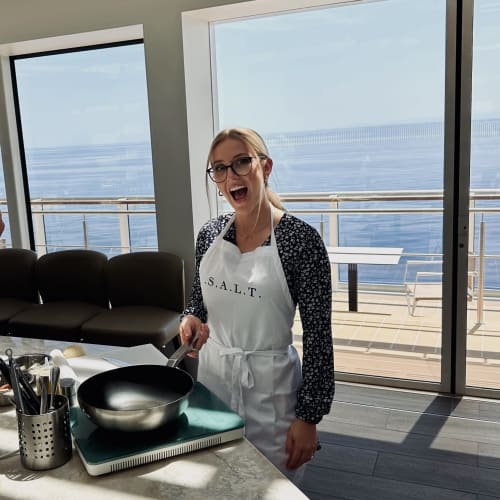 Eine Frau mit Brille und weißer Schürze mit der Aufschrift 'S.A.L.T.' steht in einem modernen Küchenbereich auf einem Schiff und hält eine Bratpfanne, hinter ihr ist durch große Fenster Meer zu sehen.