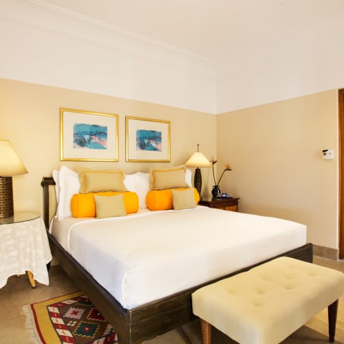 Ein luxuriöses Hotelzimmer mit einem großen Bett, dekoriert mit gelben und orange Kissen, flankiert von Tischlampen, mit eingerahmten Kunstwerken über dem Bett und einer Bank am Fuß des Bettes in einem gemütlichen, gut beleuchteten Raum.