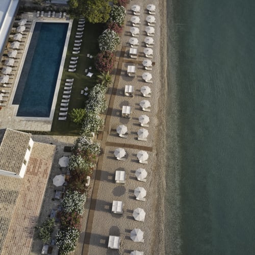 Aerial view of a coastal resort with a swimming pool, rows of white umbrellas and lounge chairs along a sandy beach beside a calm sea.