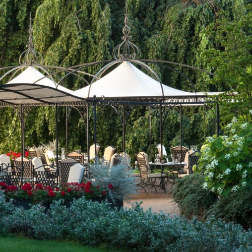 An outdoor restaurant terrace with white canopy roofs, surrounded by lush greenery and flowering plants.