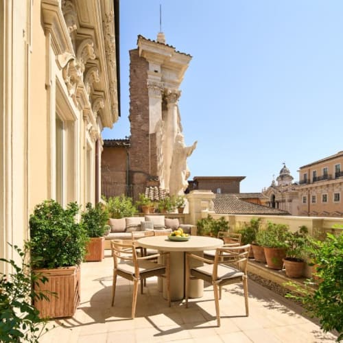 A sunlit terrace with a round table and four chairs surrounded by potted plants, overlooking historic buildings in Rome.