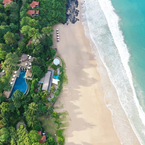 Luftaufnahme eines üppig grünen Resorts mit Schwimmbecken und mehreren Gebäuden mit roten Dächern neben einem Sandstrand mit sanften Wellen eines türkisfarbenen Meeres.