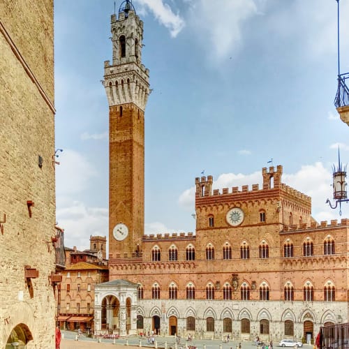 Palazzo Pubblico with the Torre del Mangia tower in Siena, Italy, during daytime with people and cars in the foreground.