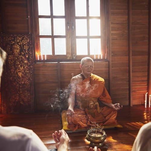 Ein Mönch führt eine Meditationssitzung mit zwei Teilnehmern in einem Holzraum auf einem Elefantenlager in Thailand an.