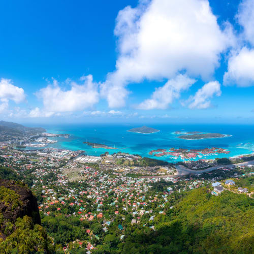 A panoramic bird's-eye view of Victoria Mahe showcasing a vibrant coastal town, lush green hills, and the blue ocean with small islands in the background under a partly cloudy sky.