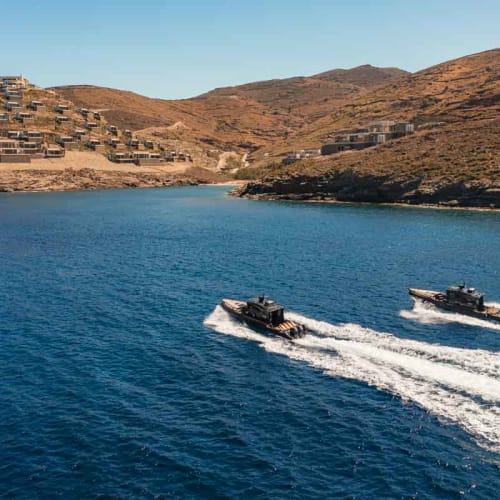 Two boats speeding on blue sea water near a rocky coastline with brown hills and scattered buildings under a clear sky.