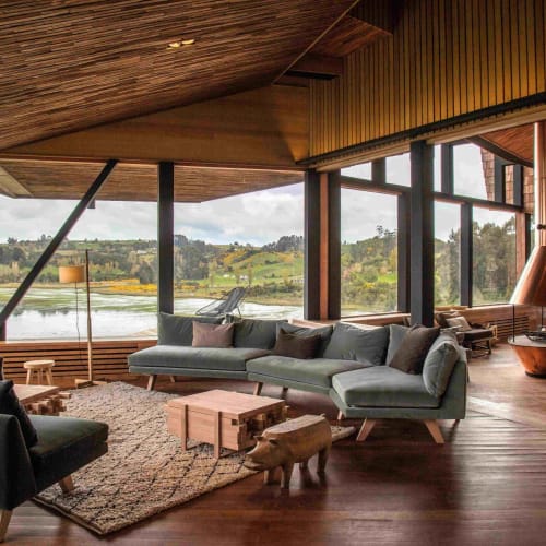 Modern living room with large windows overlooking a lake and green landscape, featuring contemporary furniture and wooden accents.