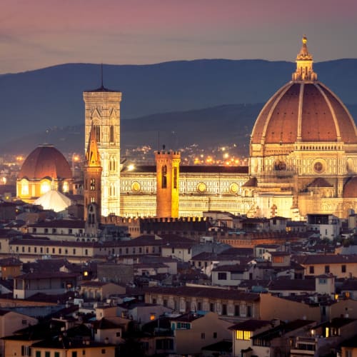 A nighttime view of Florence featuring the illuminated Florence Cathedral with its prominent dome, surrounded by city buildings and distant mountains.