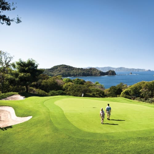 Two people walk on a lush green golf course overlooking the ocean with trees and hills in the background.