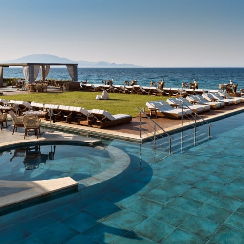 A luxurious outdoor pool area with lounge chairs and a shaded dining pavilion overlooking the sea on a clear day.
