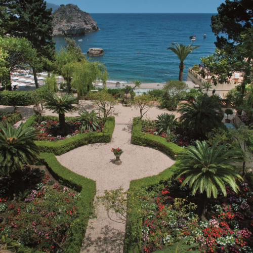 Ein üppiger Garten mit symmetrischen Blumenbeeten, großen Palmen und einem Weg, der zu einem Strand mit Felsen und dem Meer im Hintergrund führt.