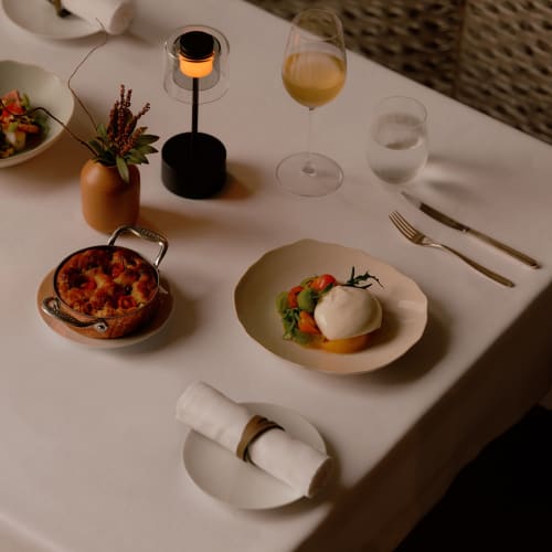 A beautifully set dinner table with a candle, glass of white wine, water, a bowl of berries, and a plate with a main course surrounded by cutlery.
