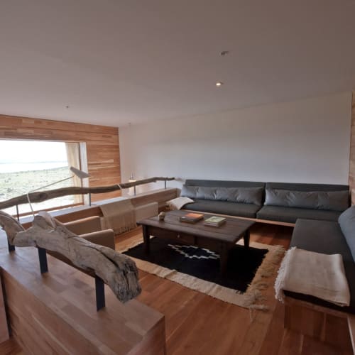 Innenansicht eines gemütlichen Wohnzimmers mit Holzakzenten, großem grauen Sofa und Logs als Dekor in Tierra Patagonia Suite, Chile.