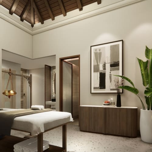 Ein ruhiger Ayurveda-Spa-Raum mit einer Massageliege mit Handtüchern, warmem Licht, Holzmöbeln, einer großen Topfpflanze und entspannender Dekoration mit gerahmter Kunst und Jalousien an den Fenstern.