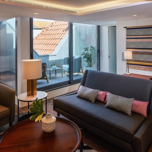 Ein gemütlicher Hotel-Wohnbereich mit einem schwarzen Sofa, Sessel und einem runden Holztisch mit Vase, in der Nähe großer Fenster mit Balkonblick.