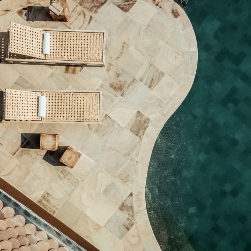 Draufsicht auf einen Swimmingpool mit geschwungener Kante neben einer Terrasse mit Steinfliesen, zwei geflochtenen Liegestühlen und Topfpflanzen.