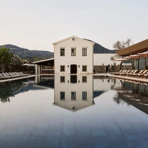 Ein ruhiger Außenpool, der ein weißes Gebäude mit Bergen im Hintergrund spiegelt, umgeben von Liegestühlen und Sonnenschirmen in einem Resort.