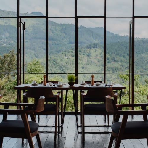 A restaurant interior with several tables and chairs facing a large window overlooking a lush, mountainous landscape.