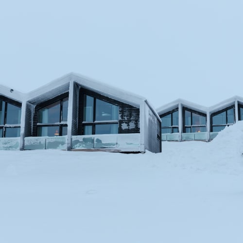 Modern Arctic accommodation with large glass windows covered in snow, surrounded by a vast snowy landscape under an overcast sky.