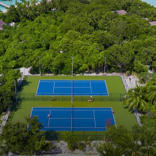 Zwei Tennisplätze in einer grünen, waldreichen Umgebung, umgeben von Bäumen und Villas, mit Blick auf den Strand im Hintergrund.