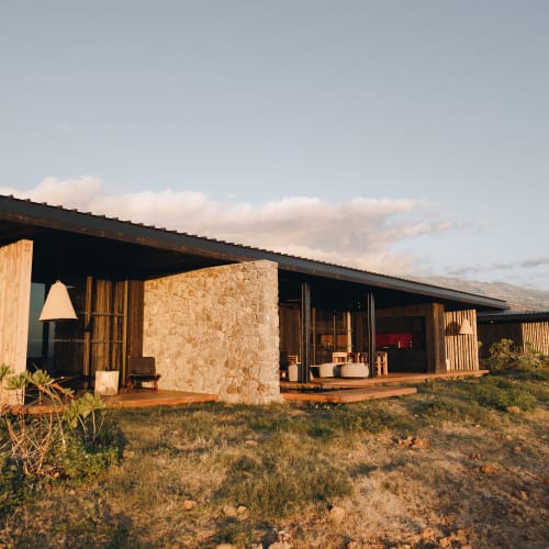 Modern single-story house with a slanted roof and stone and wood walls, situated in a dry grassy landscape under a clear sky.