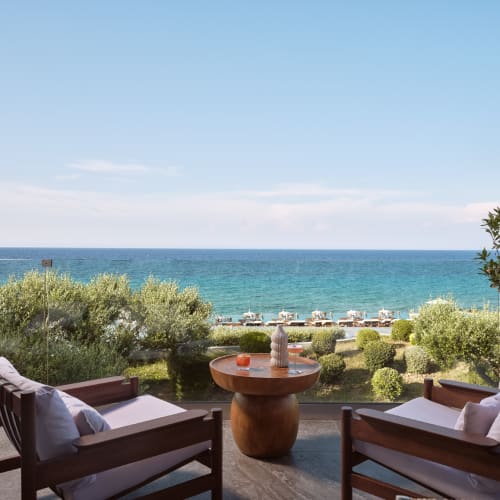 Two wooden chairs with cushions and a small round table with drinks, set on a terrace overlooking a green garden and the blue sea under a clear sky.