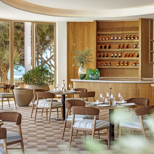 Elegant restaurant interior with large windows offering ocean view, wooden shelves with bottles, and nicely set tables with white tableware.