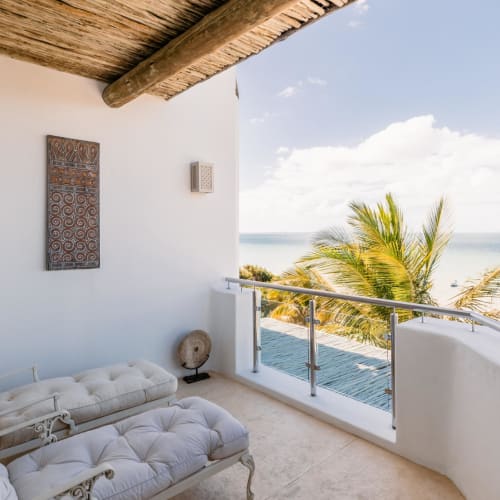 A luxurious balcony with a white chaise lounge overlooking the ocean, palm trees, and a partly cloudy sky.