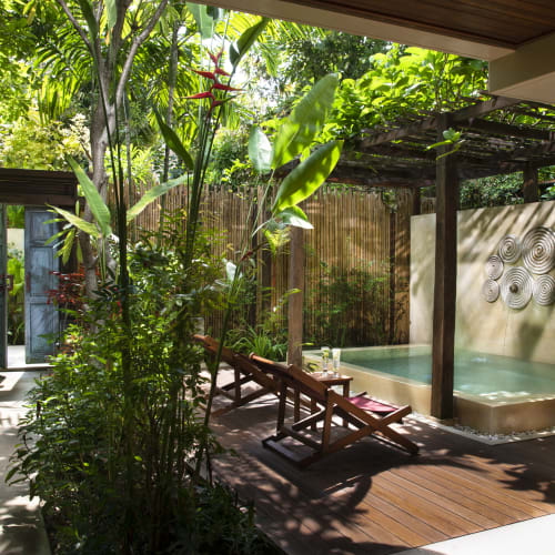 A shaded outdoor tropical patio with two wooden lounge chairs and a small private pool surrounded by lush green plants and wooden fencing.